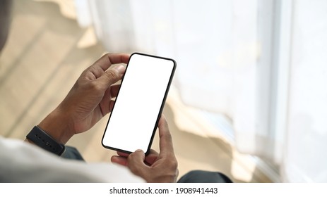 Close Up View Of Young Man Sitting Near Window And Holding Mock Up Smart Phone With White Screen.