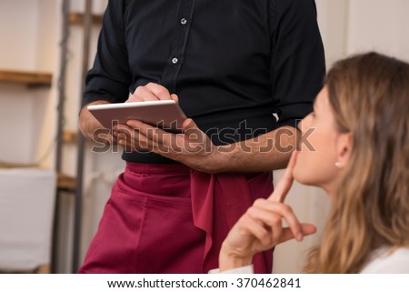 Close up of waiter hand noting down menu on tablet. Young woman ordering for food to a waiter at restaurant. Young beautiful woman thinking of food to order in front of a waiter holding tablet. 