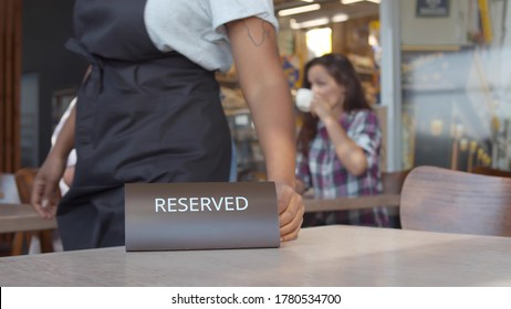 Close Up Of Waiter Putting Reserved Plate On Table In Restaurant With Happy Couple Drinking Coffee On Date On Blurred Background. Restaurant Manager Placing Reserved Table Sign