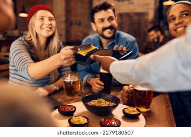 Close up of waitress holding credit card reader while customers are making contactless payment in a bar.