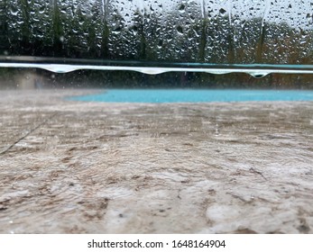 Close Up Of Water Dripping From A Glass Swimming Pool Fence With A Pool In The Background And Rain Falling Onto The Stone Tiles And Overflowing Puddles.