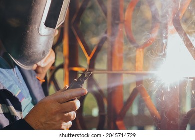 Close Up Of A Welder Hands Welding Steel Structure Metal Part In Home Backyard. Craft Man Use Electric Arc Argon Wear Work Uniform,protective Helmet On Head Without Gloves While Sparks Fly At Factory.