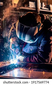 Close Up Of A Welder Welding In Workshop. He Is Dressed In A Work Uniform And Has A Protective Helmet On His Head While Sparks Fly In All Directions.