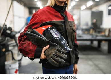 Close up of welding mask in hands of woman welder in workshop. Female worker operating welding machine, wearing protective clothing and a welding mask. - Powered by Shutterstock