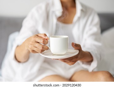 Close up of white ceramic cup with hot aroma coffee in female hands. Mature lady in sleepwear drinking morning beverage while sitting on bed.