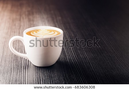 Close up white coffee cup with heart shape latte art foam on black wood table near window with light shade on tabletop at cafe.