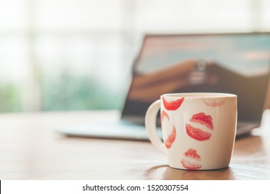 Close up of white cup of hot coffee with mark lips on the top. Mug of coffee with red lipstick marks on wooden table next to blurry background of laptop. Horizontal shot - Powered by Shutterstock