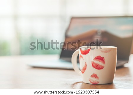 Close up of white cup of hot coffee with mark lips on the top. Mug of coffee with red lipstick marks on wooden table next to blurry background of laptop. Horizontal shot