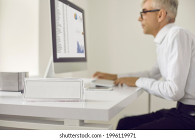 Close Up Of White Empty Name Plate In Office On Desk On Background Of Man Working On Laptop. Free Space For Writing The Name Of The Department, Position, Name Of The Head Or Employee.