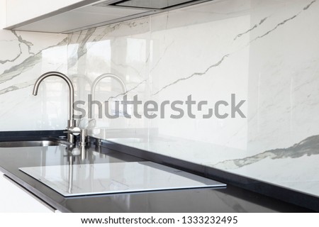 Close up of white glossy kitchen with black quartz countertop and marble tile backsplash. Build-in hidden incorporated hood and undermounted sink. 