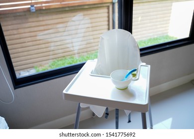 Close Up White High Chair Next To The Window In House, A Empty White Bowl With Blue Spoon Are Prepared For Infant Meal ,Baby Feeding Concept. 