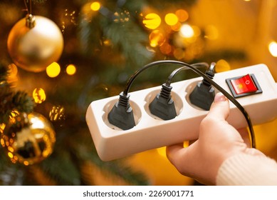 Close up of white multiple electrical socket in woman hands with three plugs in it, close up. Christmas tree with bright garland lights and golden baubles, home cozy atmosphere