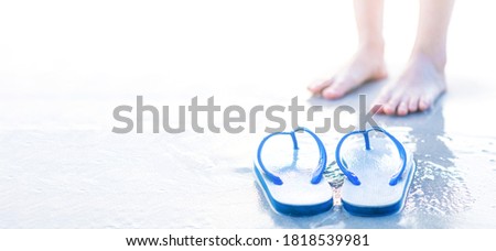 Close up of white sandals or slipper on white beach on clear day in the sun, blurry feet standing in the background. Panorama with copy space. Idea for colorful summer travel season. Selective focus.