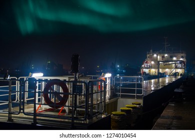Close up wide angle view of a small cargo ship docked on the terminal on a cold winter night in december with aurora borealis northern lights in the sky.