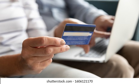 Close wife fingers holding plastic credit card on background computer on husband lap, old couple booking hotel using on-line app service and website, make online payment, shopping via internet concept