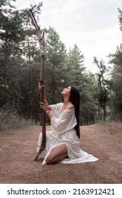 close up wild shaman. young brunette girl in white boho style dress is sitting with long high wooden witch staff in hand and looking up on a pine forest background. lifestyle concept, free space