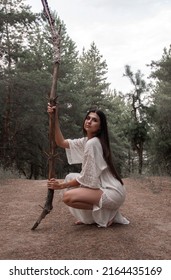 close up wild shaman. young brunette girl in white boho style dress is sitting with long high wooden witch staff in hand and looking apart on a pine forest background. lifestyle concept, free space