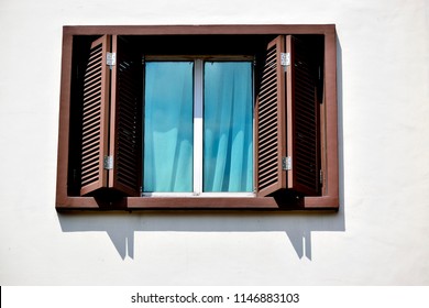 Close Up Of Window With Brown Teak Bi Fold Wooden Shutters With Blue Glass On A White Wall Of House In Singapore