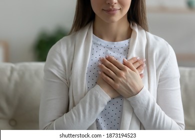 Close up wishful peaceful young woman holding hands on chest, making wish, grateful sincere girl thanking fate, meditating, praying, sitting on couch at home, showing gratitude sign