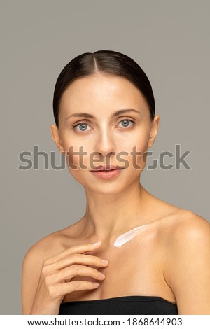 Close up of woman applying sunscreen cream or cosmetic moisturizing lotion on perfect hydrated soft skin on chest, grey background. Beauty procedure, body cream advertisement concept. 