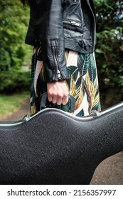 Close Up Of Woman Arm Holding A Guitar Case In A Park. She Is Wearing A Black Leather Jacket And Spring Dress. Vertical.