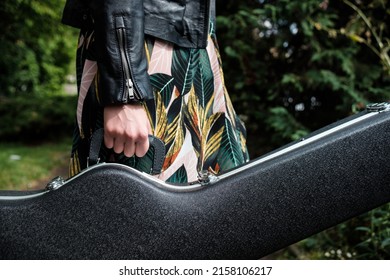 Close Up Of Woman Arm Holding A Guitar Case In A Park. She Is Wearing A Black Leather Jacket And A Spring Dress. Horizontal.