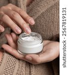 Close up of woman in brown bathrobe touching cream with finger and holding opened cosmetic jar in other hand closeup, natural manicure. Organic cosmetics for body and face care