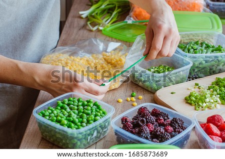 Close up of woman closing plastic storage bag with fresh corn for freezing on the wooden table. Plastic food boxes with another fresh vegetables and berries around it.