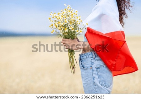 Close up of woman covered with  flag of Poland holding bouquet of daisies in wheat field. Polish Flag Day. Independence Day. Travel and learn polish language concept.