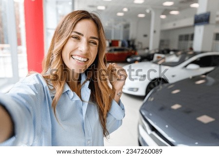 Close up woman customer buyer client in shirt do selfie shot pov on mobile phone do winner gesture choose auto want buy new automobile in car showroom vehicle salon dealership store motor show indoor.