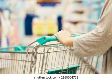 Close Up Woman With Empty Green Shopping Cart At Supermarket Aisle. Female And Trolley Cart At The Product Shelf In The Supermarket. Grocery Store With Many Food And Appliances In Modern Trade Hall.