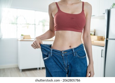 Close Up Of Woman Feel Happy After Lose Weight For Health In House. Attractive Beautiful Skinny Girl Wearing Old Big Jeans With Abs Abdomen Sick Packs In Kitchen At Home. Healthy Food And Diet Concept