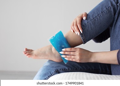 Close Up Woman Feet And Legs, And Hands Holding Ice Gel Pack As Cold Compress On Ankle Due To Stretching Or Injury, Sitting On Bed In Apartment Indoors