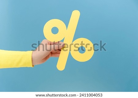 Close up of woman hand holding paper yellow percent mark, presenting big discounts, posing isolated over plain blue color background wall in studio with copy space for advertisement
