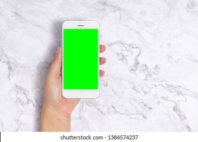 Close Up Woman Hand Holding White Mobile Phone With Blank Green Screen On White Marble Background, Front View With Copy Space. Clipping Path Include