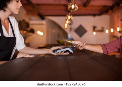 Close Up Of Woman Hand Paying With Smart Phone Using NFC Technology. Paying With Just A Tap In A Coffee Shop During Daytime. Concept Business, Technology, Contactless.