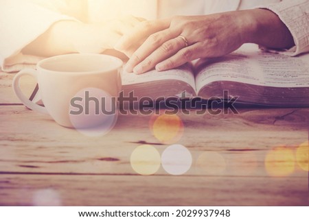 Close up of a woman hand  prays while reading the open bible, blurred page with a coffee cup on wooden table with window light , Christian prayers or bible study concept background with copy space