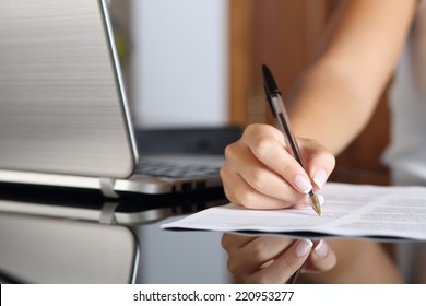 Close Up Of A Woman Hand Writing A Contract With A Laptop Beside At Home Or Office