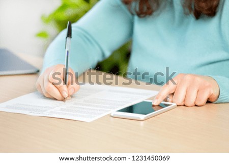 Close up of a woman hand writing or signing in a document on consulting a mobile phone on a desk at home or office