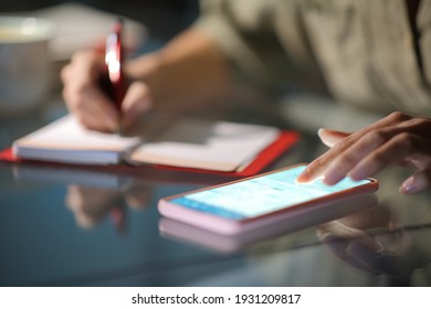 Close up of a woman hands checking smart phone calendar and writing on agenda in the night at home
