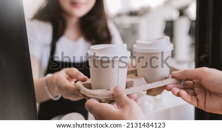Close up woman hands gives paper coffee cup on slide glass window. Take away delivery concept. Asian woman barista. Cropped shot of waitress and buyer hold coffee, eco recycle environment friendly.