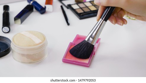 Close Up Of Woman Hands Holding Makeup Brush Applying Pink Blush On White Vanity Table. Female Using Cosmetics Products For Make Up. Messy Dressing Table With Power And Palette Indoors.