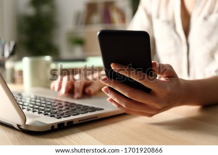 Close up of woman hands using laptop checking smart phone at night at home
