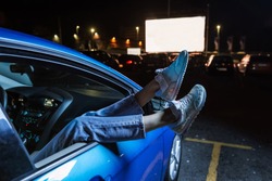 Close Up Of Woman Hanging Her Legs Out Of Car Window While Watching A Movie At Drive In Cinema From The Front Seat Of The Car. Concept Of Carefree Leisure Time