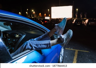 Close up of woman hanging her legs out of car window while watching a movie at drive in cinema from the front seat of the car. Concept of carefree leisure time