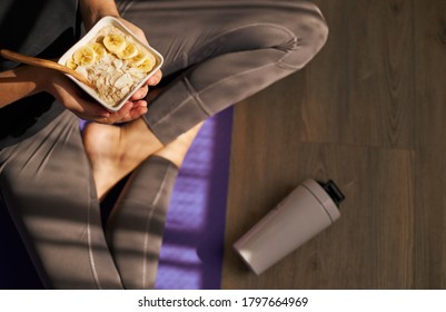 Close up of woman holding healthy breakfast bowl sitting on yoga mat after workout.