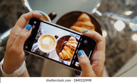 Close up of woman holds modern smartphone and taking pictures of her morning breakfast coffee and tasty croissant buns to share photos on social media resources