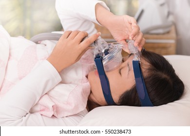 Close Up Of Woman Laying On Back Holding And Adjusting Cpap Mask  In Bed With Cpap Machine In Background, Side View.Obstructive Sleep Apnea Therapy. 