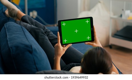 Close up of woman looking at tablet computer in horizontal mode with mock up green screen chroma key display lying on couch. Freelancer using isolated touchscreen device for social networks browsing