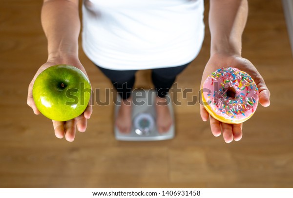 Close up of woman on scale holding on hands\
apple and doughnut making choice between healthy unhealthy food\
dessert while measuring body weight in Nutrition Health care Diet\
and temptation concept.